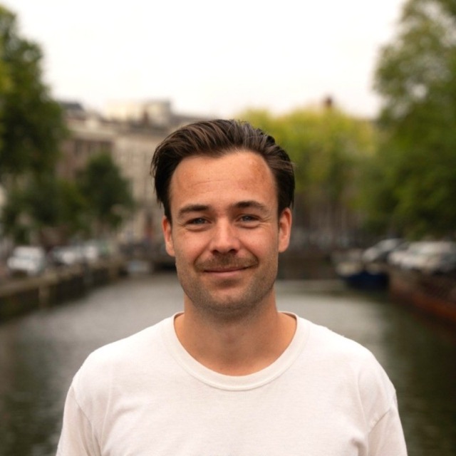 Thomas Lock, glimlachend in een wit T-shirt langs een Amsterdamse gracht